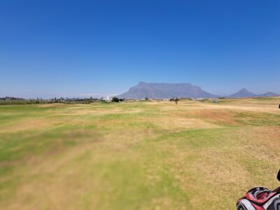 Wide-looking fairways at Milnerton Golf Club running alongside each other, with a mountain rising in the background and a small lighthouse visible to the left.