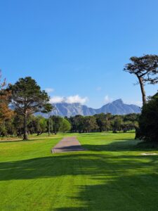 Early morning at Royal Cape Golf Club with bright skies and a mountain backdrop partially covered by a few clouds, dew on the grass, wheel tracks across the fairway, and a pathway running along the side.