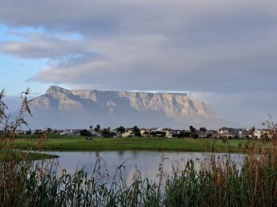 Gloomy sky over Milnerton Golf Club with grey cloud cover, a water hazard showing a faint reflection of the nearby mountain, and houses on the estate visible in the background.