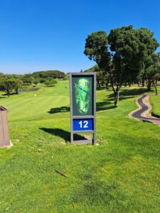 Tee box at the 12th hole of Rondebosch Golf Club showing a course map sign indicating the fairway layout, with the fairway stretching ahead, a cart path running alongside it, and trees lining the edges.