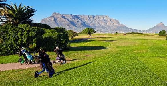 Three golf trolleys lined up on the left side near shrubs at Milnerton Golf Club, with the fairway stretching ahead under clear skies and a mountain in the background.